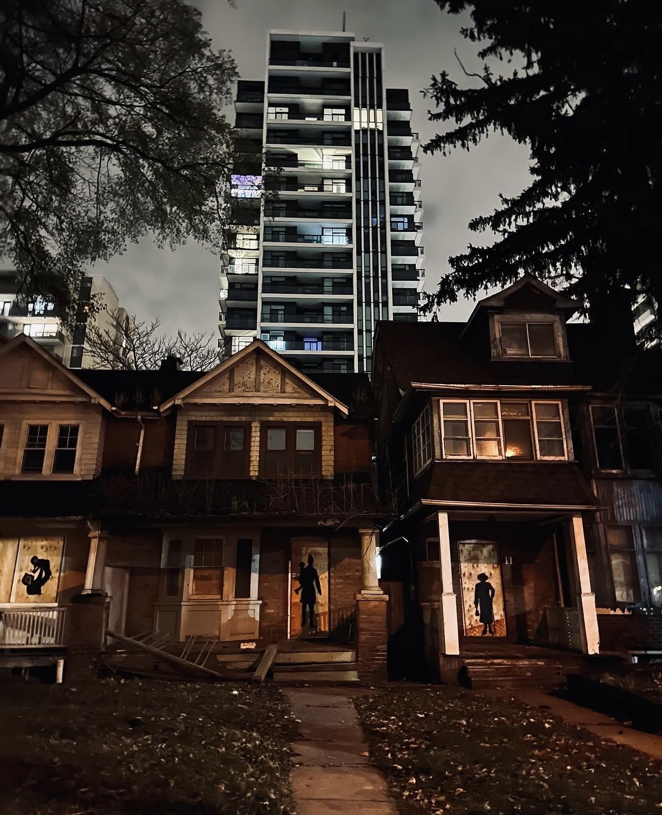 Derelict row houses at night with silhouette art in doorways and condo tower behind
